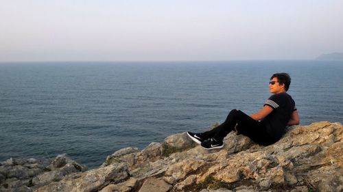 Young woman sitting on retaining wall by sea against clear sky