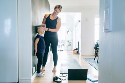 Smiling mother measuring daughter's height on wall in living room at home