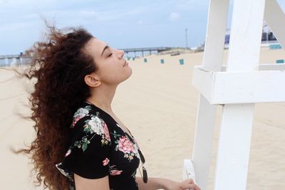 Side view of young woman with long curly hair standing at beach against sky