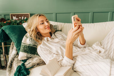 Young attractive blond woman lying on the sofa uses a mobile phone to communicate.