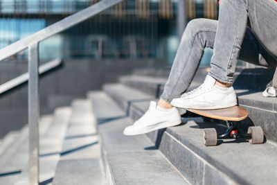 Man in grey jeans and white sneakers put his feet on the longboard. hipster sitting on the steps