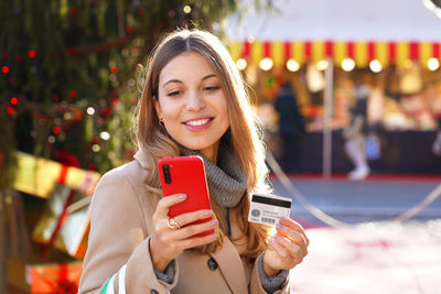 Woman paying her gifts with smartphone and credit card with christmas markets on the background