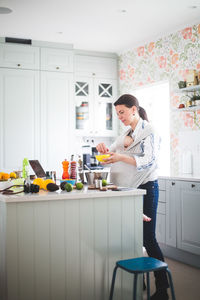 Working mother blogging in kitchen while carrying daughter in baby carrier at home