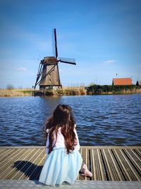 Rear view of girl sitting on pier by lake against sky