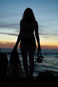 Rear view of silhouette woman standing at beach during sunset