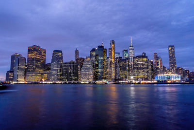 Illuminated modern buildings by river against cloudy sky at dusk