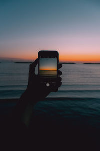 Person holding smart phone by sea against sky during sunset