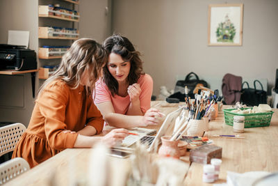 Two young female entrepreneurs sit with laptop in their workshop