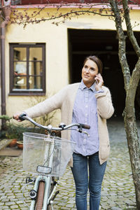 Smiling businesswoman using mobile phone while standing with bicycle on sidewalk