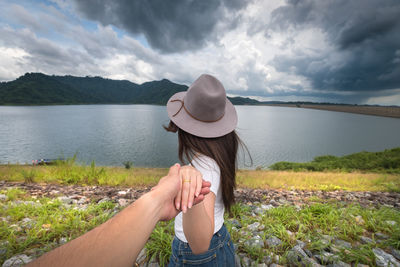 Cropped image of man holding women hand at riverbank against cloudy sky