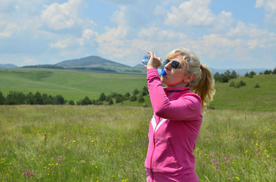 Woman dressed in sport clothes drinking water from a plastic bottle in a picturesque landscape.