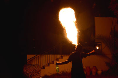 Rear view of man blowing fire against building at night