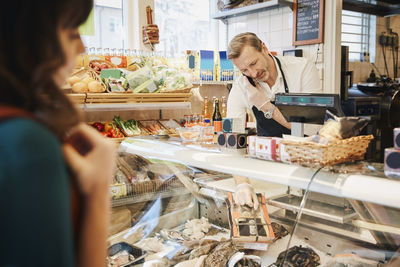 Male owner talking on mobile phone while picking up product from display cabinet for customer in store