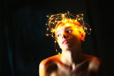 Close-up of shirtless teenage boy with illuminated string lights on head against black background