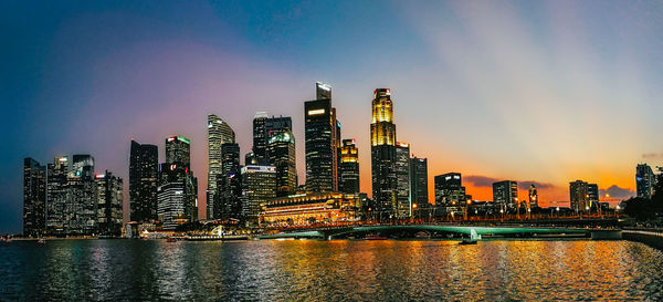 Illuminated modern buildings by river against sky in city