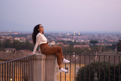 Side view of woman sitting on railing against cityscape