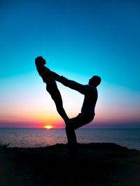 Silhouette couple exercising at beach against sky during sunset