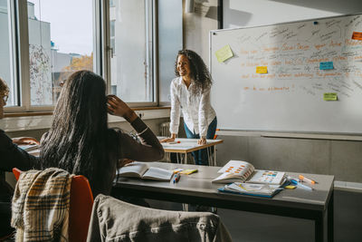 Cheerful professor teaching university students while leaning on desk in classroom