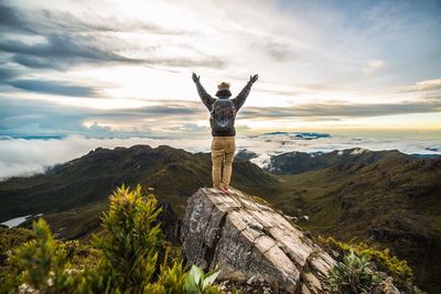 Full length rear view of man with arms raised standing on rock at mountain against sky
