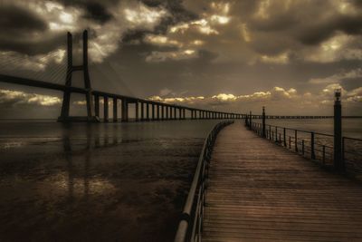 Empty boardwalk and bridge over sea against cloudy sky during sunset