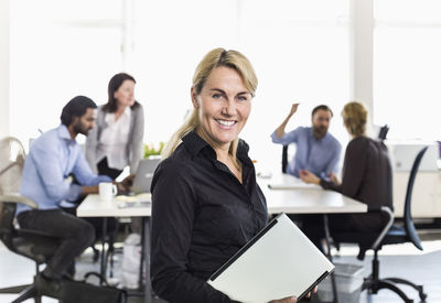 Portrait of mature businesswoman holding laptop with colleagues in background at office