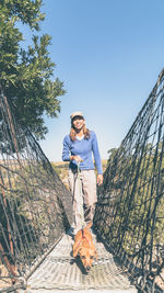 Smiling woman walking with dog on footbridge against clear blue sky