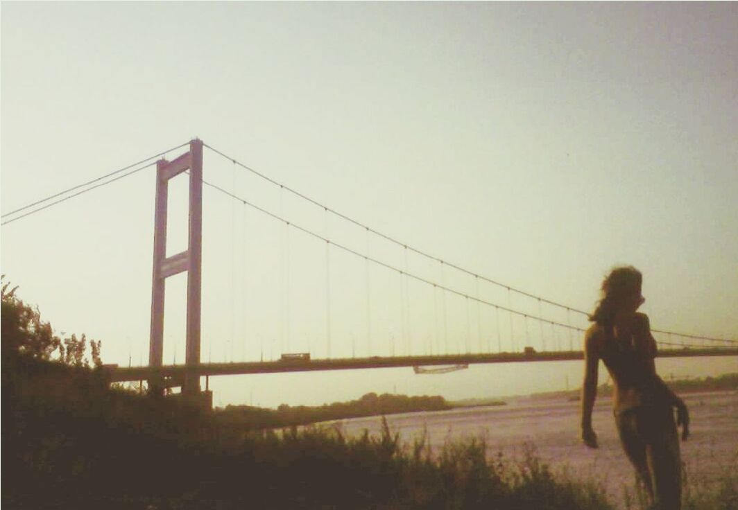 clear sky, lifestyles, copy space, leisure activity, water, built structure, standing, full length, architecture, rear view, connection, men, bridge - man made structure, side view, sea, person, sky, casual clothing