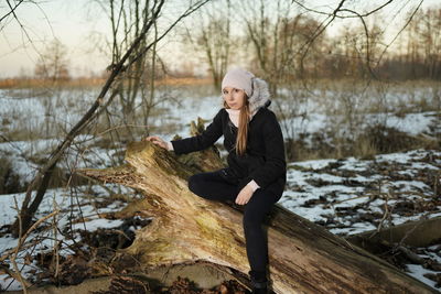 Full length of woman sitting by tree trunk outdoors during winter