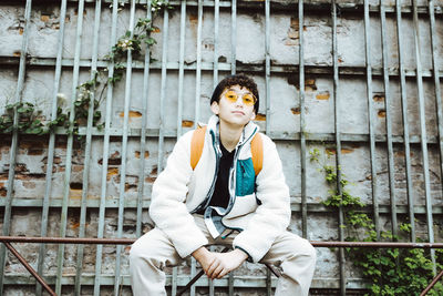 Portrait of boy wearing sunglasses sitting on railing against wall during weekend