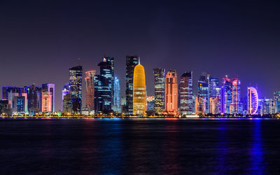 Illuminated doha tower and skyscrapers by river in city against clear sky at night