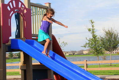 Cheerful girl sliding on slide at playground against sky