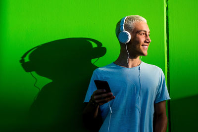 Smiling teenage boy listening music while standing against green wall