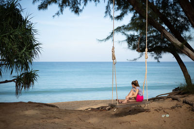 A girl sits on a swing on the beach and looks at the sea hugging her knees