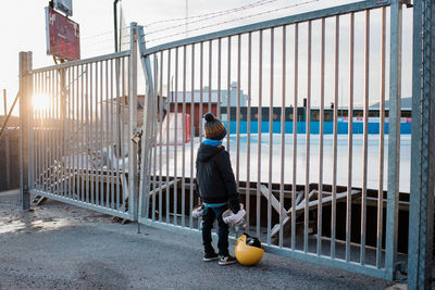 Boy holding skates looking through a fence at an empty ice rink