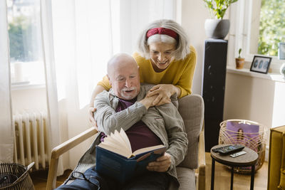 Happy senior woman with arm around man reading book while sitting on chair at home