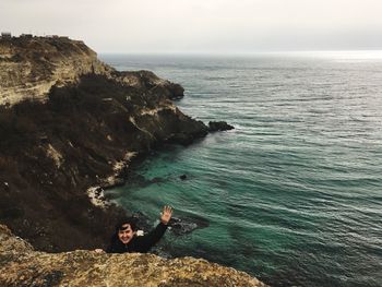 Portrait of smiling man gesturing while climbing on cliff against sea