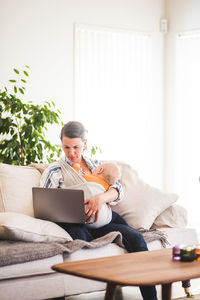 Multi-tasking mother using laptop while caring baby girl on sofa at home office