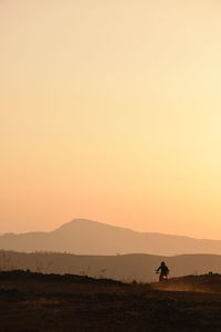 Silhouette man riding motorcycle on landscape against clear orange sky