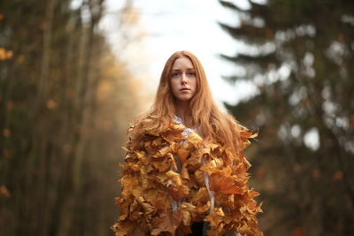 Portrait of young woman covered with leaves standing at forest during autumn