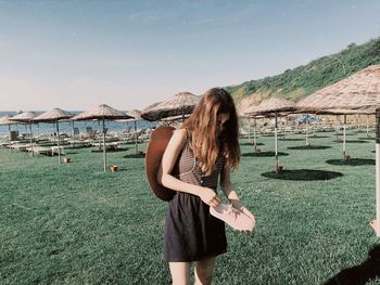 Woman holding shoe while standing on grassy field against sea