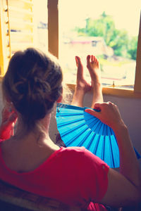 Rear view of woman holding blue folding fan while sitting at home