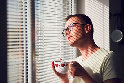 Close-up of young man having coffee while looking through window at home