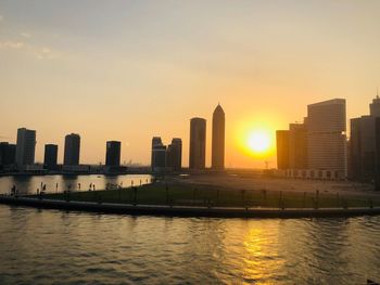 Modern buildings by river against sky during sunset