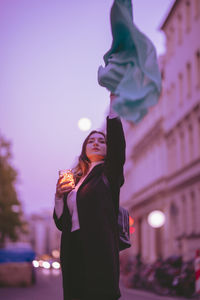 Beautiful woman moving green scarf while holding illuminated jar in city at dusk