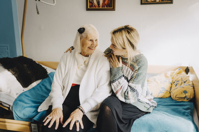 Female caregiver talking with elderly woman sitting on bed at retirement home