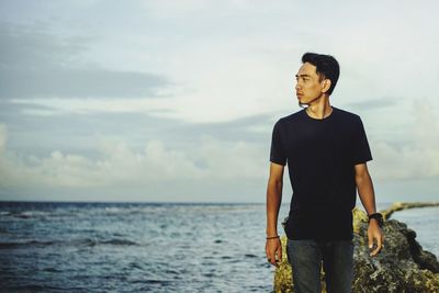 Young man looking away while standing by rock against sky