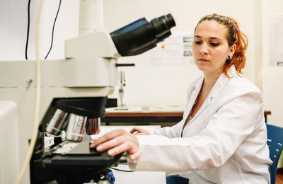 Focused female scientist in medical white uniform putting chemical sample into modern microscope while working at table in light laboratory