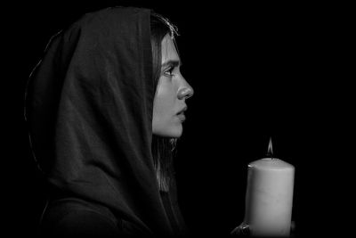 Thoughtful young woman with candle wearing hood against black background