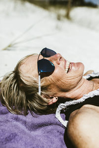 Cheerful mature woman wearing sunglasses while lying at beach during sunny day