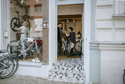 Male owner repairing bicycle of senior customer while colleagues working at workshop seen through doorway